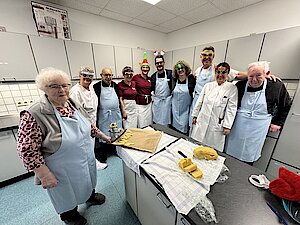 Eine Gruppe älterer Menschen mit Schürzen stehen nebeinander in einer großen Küche. Manche tragen weihnachtliche Brillen.